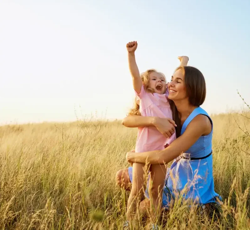 Eine Mutter umarmt ihr fröhliches Kind in einem Feld unter blauem Himmel.