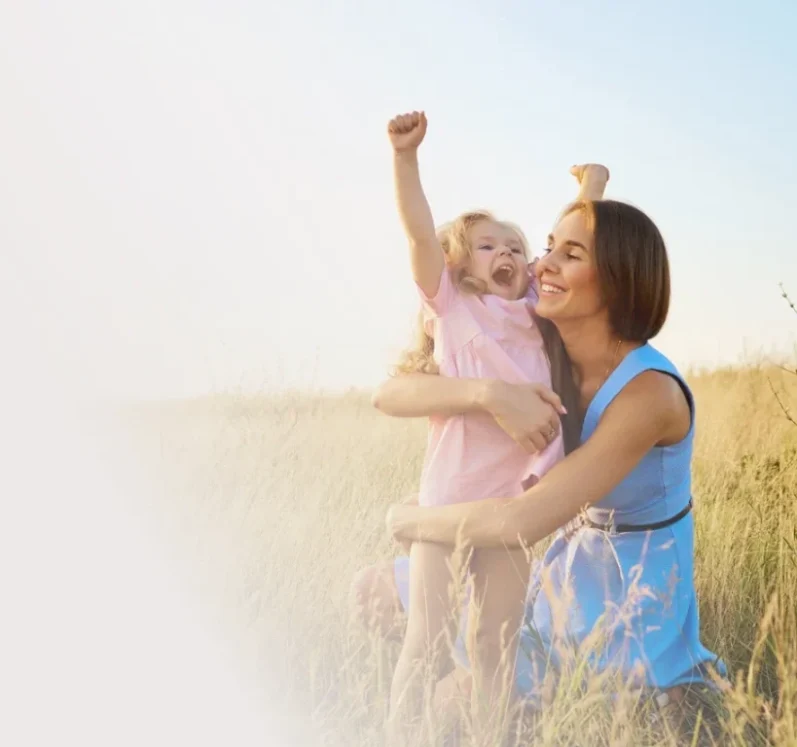 Eine Mutter umarmt ihr fröhliches Kind in einem Feld unter blauem Himmel.