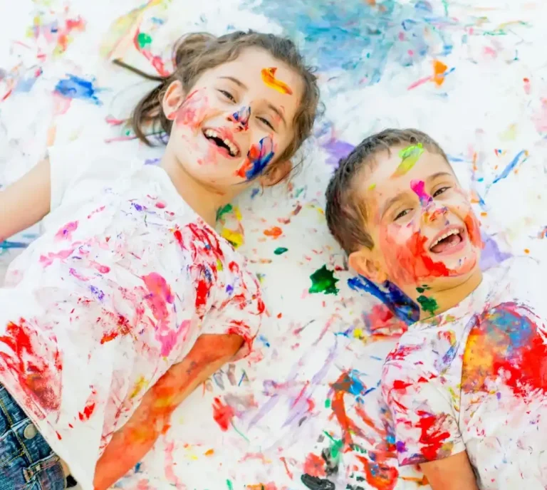 Zwei fröhliche Kinder, die mit Farbe spielen und lachend auf einem weißen Untergrund liegen.
