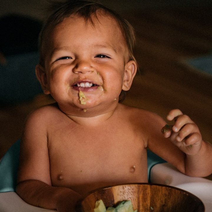 Lächelndes Kleinkind mit Essen in der Schüssel, auf dem Tisch sitzend.