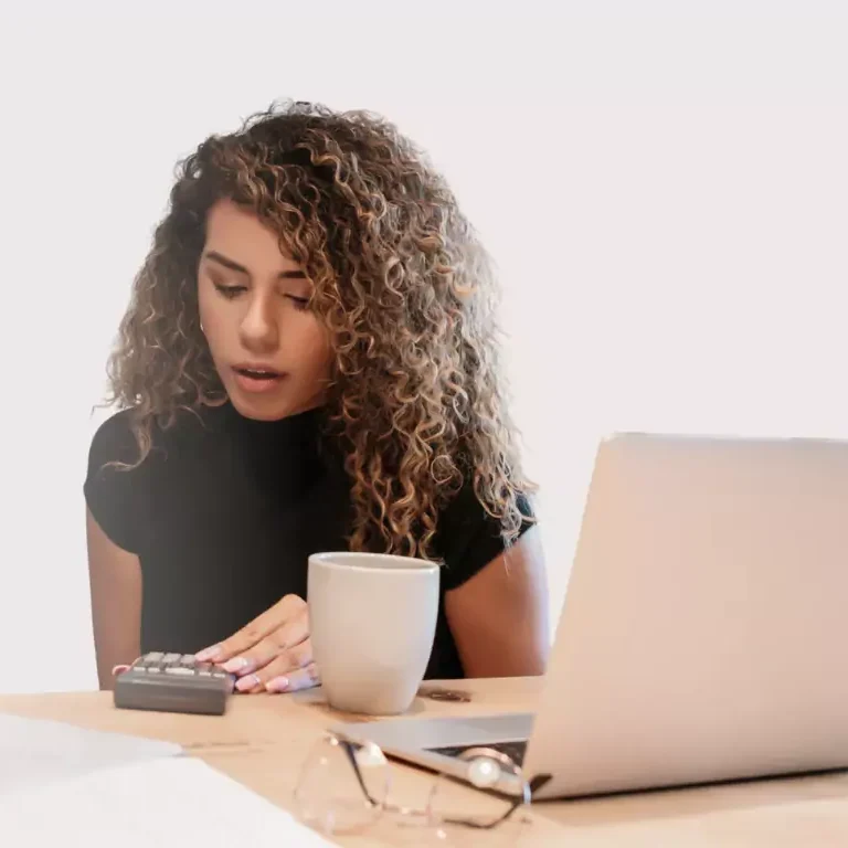 Junge Frau mit lockigem Haar, die am Tisch mit einem Taschenrechner und einer Tasse sitzt.